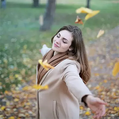 femme-jour-d-automne-feuilles-tombantes copie desktop Jeune femme aux taches de rousseur, les yeux fermés, portant un manteau beige et une écharpe marron, profite d'une journée d'automne avec des feuilles jaunes tombant autour d'elle.
