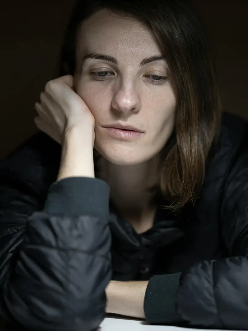 femme-pensif-bras-visage Gros plan d'une femme aux cheveux bruns, le visage appuyé sur sa main, le regard pensif et baissé, vêtue d'une veste noire.