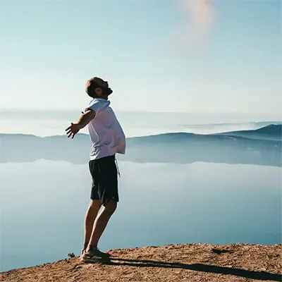 homme-bras-ouverts-sommet-montagne copie desktop Un homme aux bras écartés, les yeux levés vers le ciel, se tient sur le bord d'une falaise surplombant une mer de nuages et des montagnes lointaines.