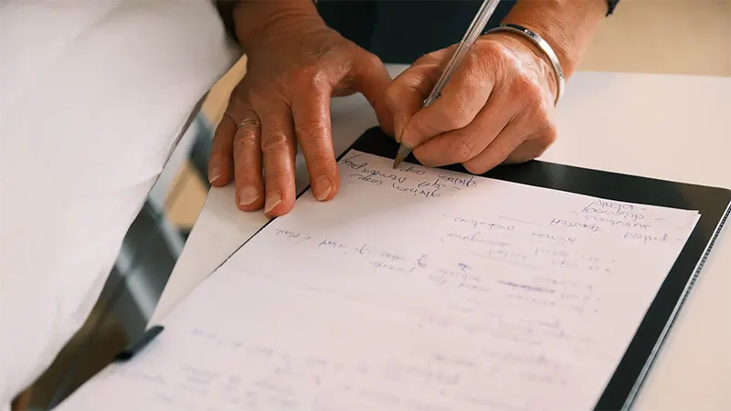 Gros plan sur les mains de Caroline Rochez kinésiologue à Uccle écrivant à la main sur un papier avec un stylo, sur un bloc-notes.