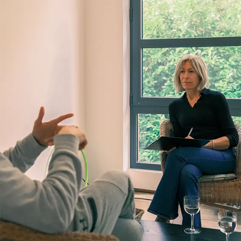 Caroline Rochez kinésiologue à Uccle assise et écrivant sur un bloc-notes, face à un patient en pleine séance, dans un cabinet lumineux.